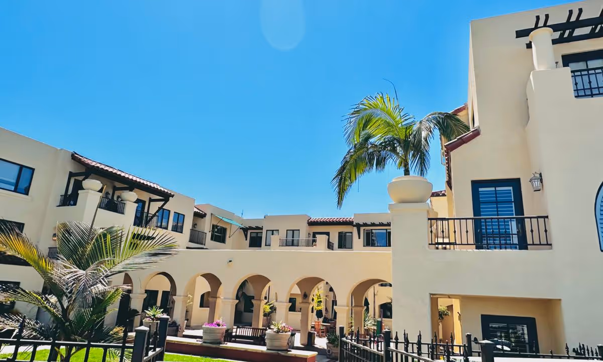 Sunlit Mediterranean-style courtyard of Casa de Mañana with arched walkways, balconies, palm trees, and potted plants under a clear blue sky.