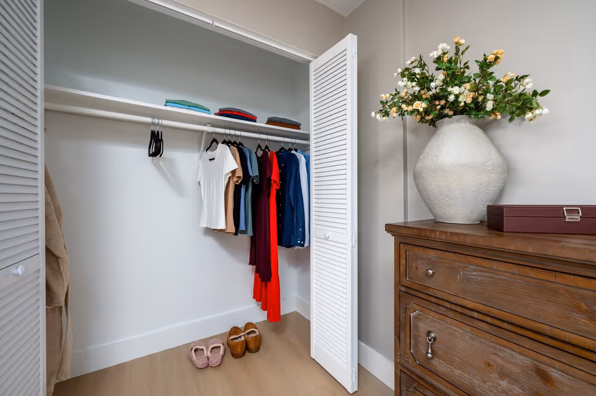 Open closet with white louvered doors containing hanging clothes including shirts and dresses, folded clothes on the top shelf, and slippers on the floor. To the right of the closet is a wooden dresser with a large white vase filled with flowers and a closed box on top.