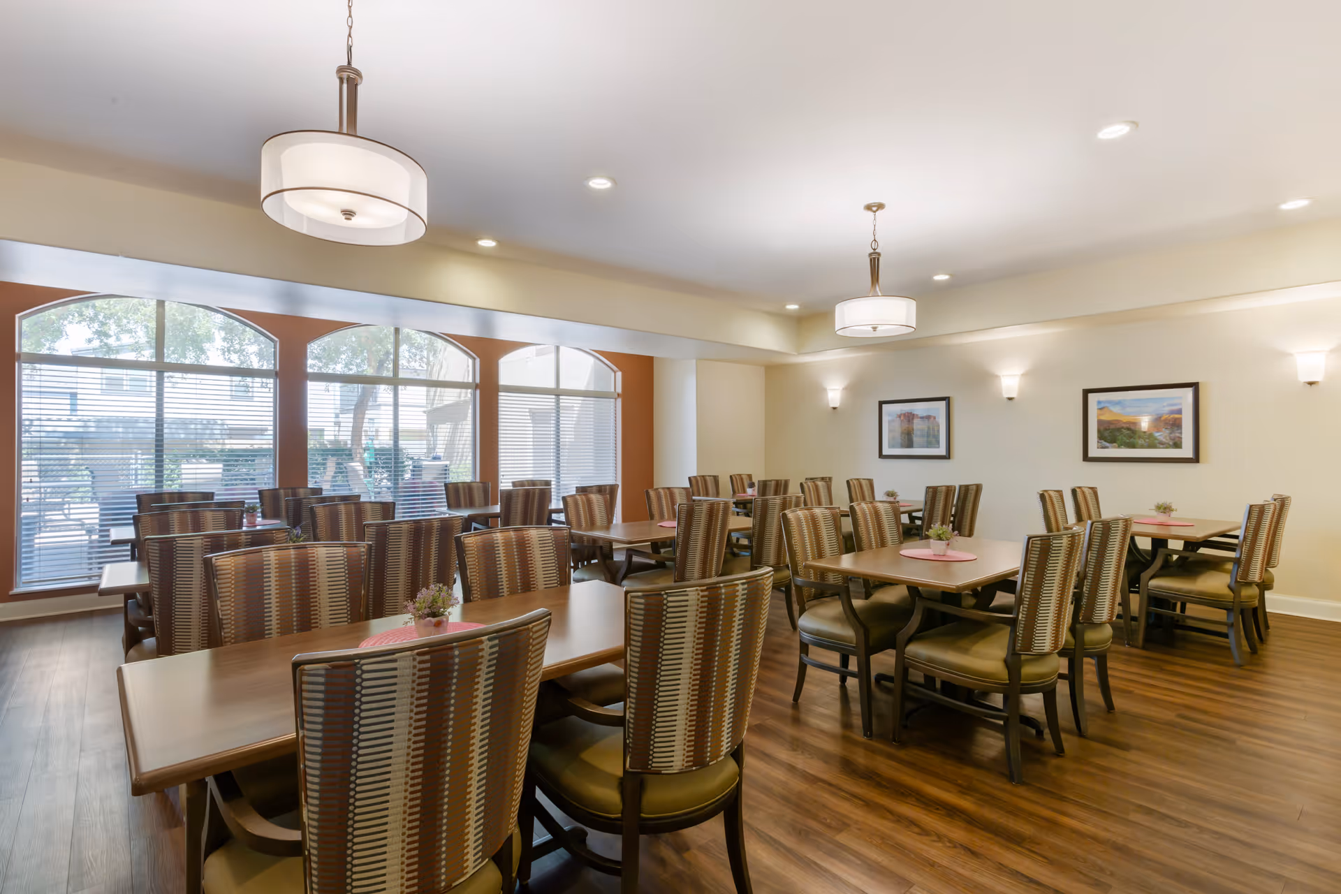 A bright dining room with multiple wooden tables and cushioned chairs arranged neatly. Large windows with blinds allow natural light to fill the room. The walls are decorated with framed landscape pictures, and modern ceiling lights hang above the tables. The floor is wooden, and small flower arrangements are placed on each table.