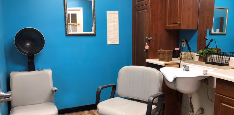 Interior of a small salon or hairdressing area with two white salon chairs, a hair dryer hood attached to the wall, a white sink with a faucet, wooden cabinets, a mirror on a bright blue wall, and various small baskets and plants on the countertop.