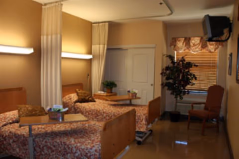 A nursing home room with two beds separated by a curtain. Each bed has a floral bedspread and a pillow. There are overbed tables with items on them, a potted plant on a small table between the beds, a window with blinds and a valance, a chair, and a wall-mounted television in the corner.
