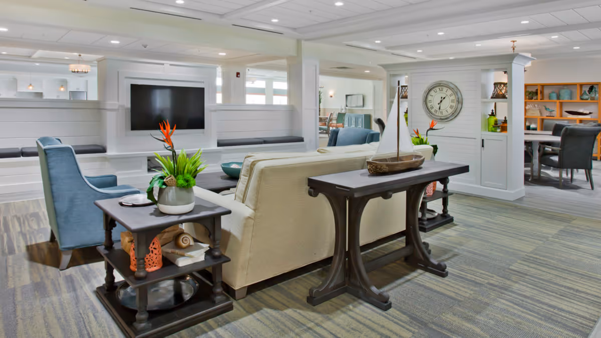 A bright and spacious living room area in a senior living facility with a beige sofa, blue armchairs, a wall-mounted flat-screen TV, decorative plants, a wooden table with a model sailboat, and a large clock on the wall. The room has a modern design with white walls, built-in shelving, and carpeted flooring.