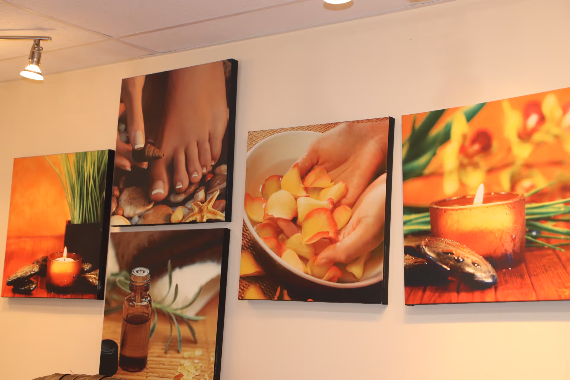 A wall with four framed pictures depicting spa and relaxation themes, including a lit candle, a close-up of feet with a seashell, hands holding rose petals in a bowl, and a bottle of essential oil with herbs.