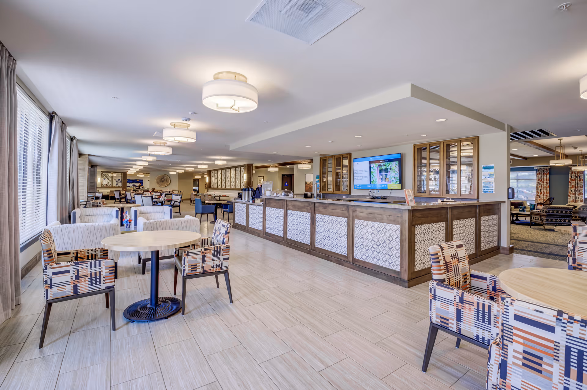 A spacious and well-lit dining area in a senior living facility with multiple tables and chairs arranged neatly. The room features patterned armchairs, round wooden tables, large windows with curtains on the left side, and modern ceiling lights. There is a long counter with decorative paneling and a large flat-screen TV mounted on the wall behind it. The area extends into a lounge space with additional seating visible in the background.