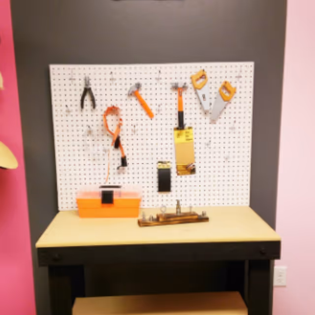 A small workbench with a pegboard mounted on the wall behind it. Various tools including pliers, a hammer, a saw, and a measuring tape are hanging on the pegboard. An orange toolbox is placed on the workbench surface. The wall behind the pegboard is painted dark gray, and the adjacent walls are pink.
