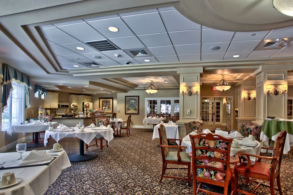 A well-lit dining room with multiple tables covered in white tablecloths, set with cups, glasses, and napkins. The room features floral upholstered chairs, patterned carpet, decorative wall sconces, and ceiling lights. Large windows with curtains allow natural light to enter, and framed artwork is visible on the walls.