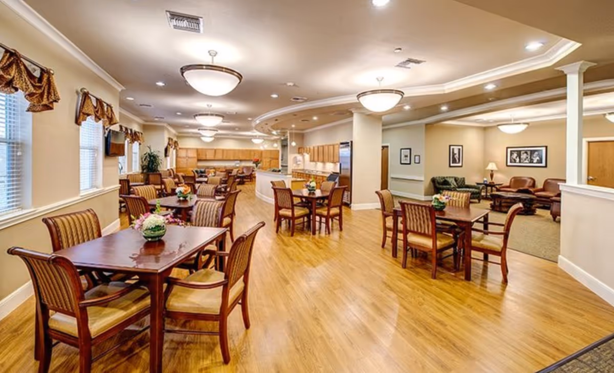 Spacious dining area with multiple wooden tables and chairs arranged neatly on a polished wooden floor. The room is well-lit with ceiling lights and has windows with decorative valances on the left wall. In the background, there is a kitchen area with wooden cabinets and stainless steel appliances. To the right, there is a cozy seating area with sofas, armchairs, a coffee table, and framed pictures on the walls.