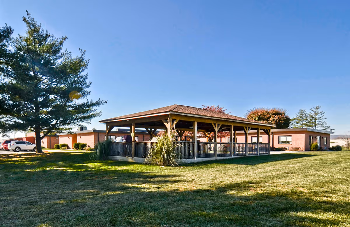 A wooden outdoor pavilion on a grassy lawn with single-story brick healthcare buildings and trees under a clear blue sky.