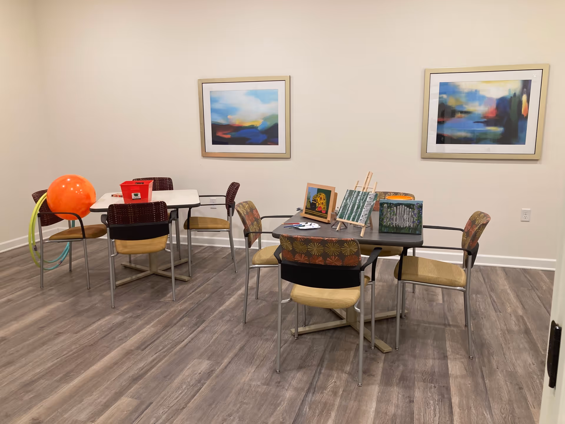 A room with two tables and several chairs arranged around them. One table has art supplies and small paintings displayed on easels. The other table has a red container, an orange exercise ball, and some hula hoops leaning against a chair. The room has light-colored walls with two abstract paintings hanging on them and wood-patterned flooring.