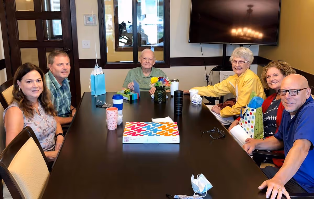 Six adults sitting around a large rectangular table in a meeting room. There are gift bags, a colorful flower arrangement, a box of Dunkin' donuts, and various cups on the table. A large TV is mounted on the wall behind them.