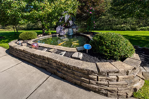 Raised stone koi pond with a small water feature, trimmed shrubs, and green lawn beside a concrete walkway.