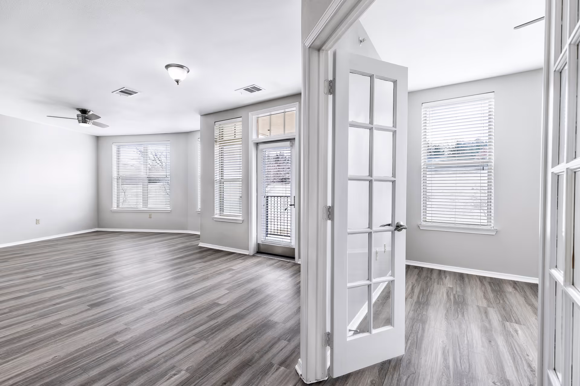 Bright and empty interior space with light gray walls and wood flooring. The room features large windows with white blinds, a ceiling fan, and a glass-paneled door leading to a smaller adjacent room with similar flooring and windows.