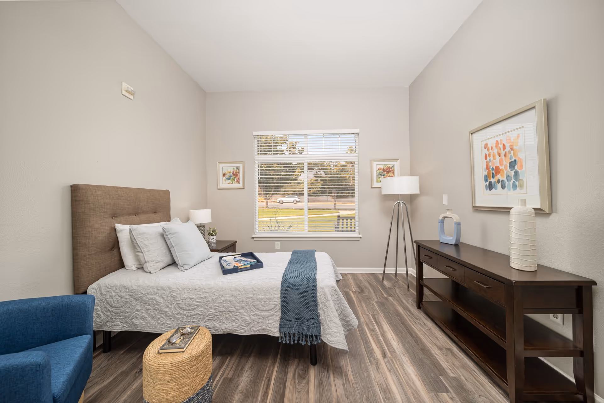 A neatly arranged bedroom with a bed featuring a brown upholstered headboard, white quilted bedding, and several pillows. A blue throw blanket is draped across the foot of the bed. Next to the bed is a dark wooden nightstand with a lamp and small plant. A blue armchair and a woven ottoman with a book and glasses are in the foreground. Across from the bed is a dark wooden console table with decorative vases and a colorful abstract painting above it. A tall floor lamp stands near the window, which shows a view of trees and a parked car outside. The room has light gray walls and wood-look flooring.