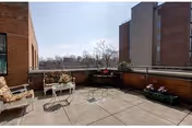 Outdoor patio area with concrete flooring, two cushioned chairs, a small white table, and flower planters along the brick walls. Surrounding buildings and a clear sky are visible in the background.
