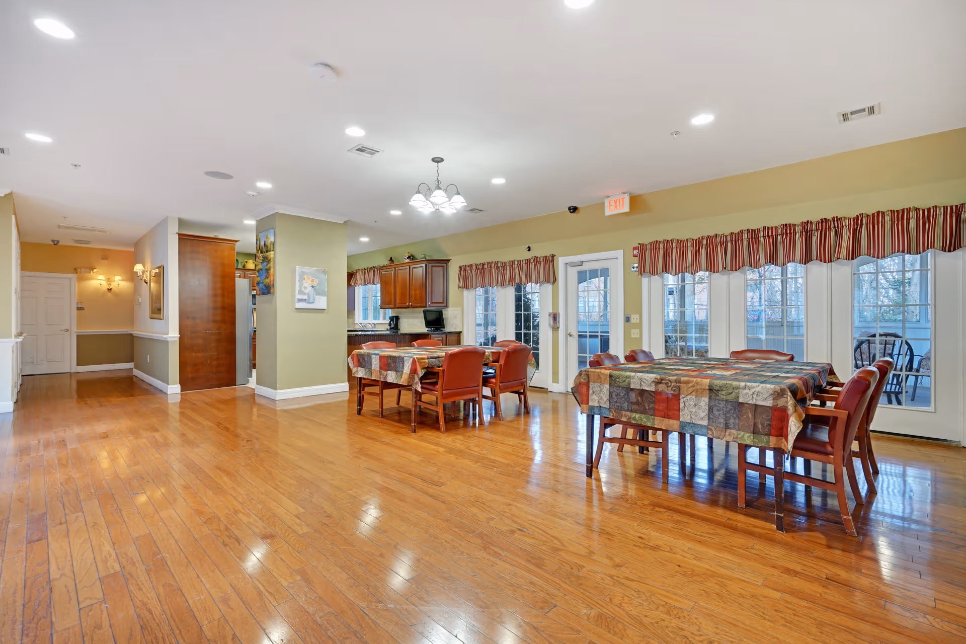A spacious dining area in a senior living facility with wooden floors, two tables covered with colorful patchwork tablecloths, and red cushioned chairs. The room has large windows with red and white striped valances, a door leading outside, and a kitchen area with wooden cabinets in the background. The walls are painted light green and beige, and ceiling lights illuminate the space.