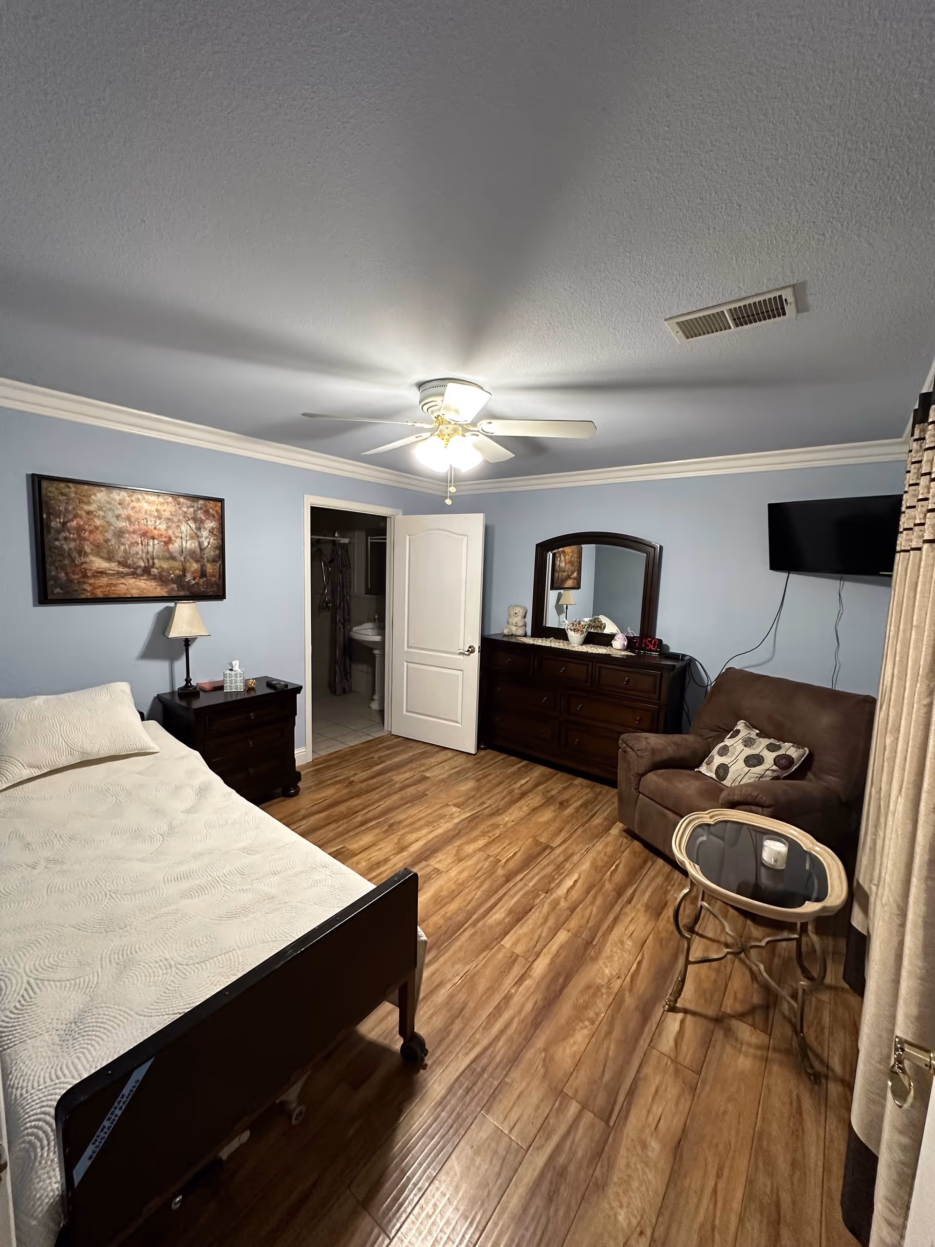 A cozy bedroom with light blue walls and wood flooring. The room features a single bed with a white quilt, a dark wooden nightstand with a lamp and tissue box, a dark wooden dresser with a mirror, a brown armchair with a patterned pillow, a small round glass-top table, and a wall-mounted TV. A door is open, revealing a bathroom with a sink and mirror.