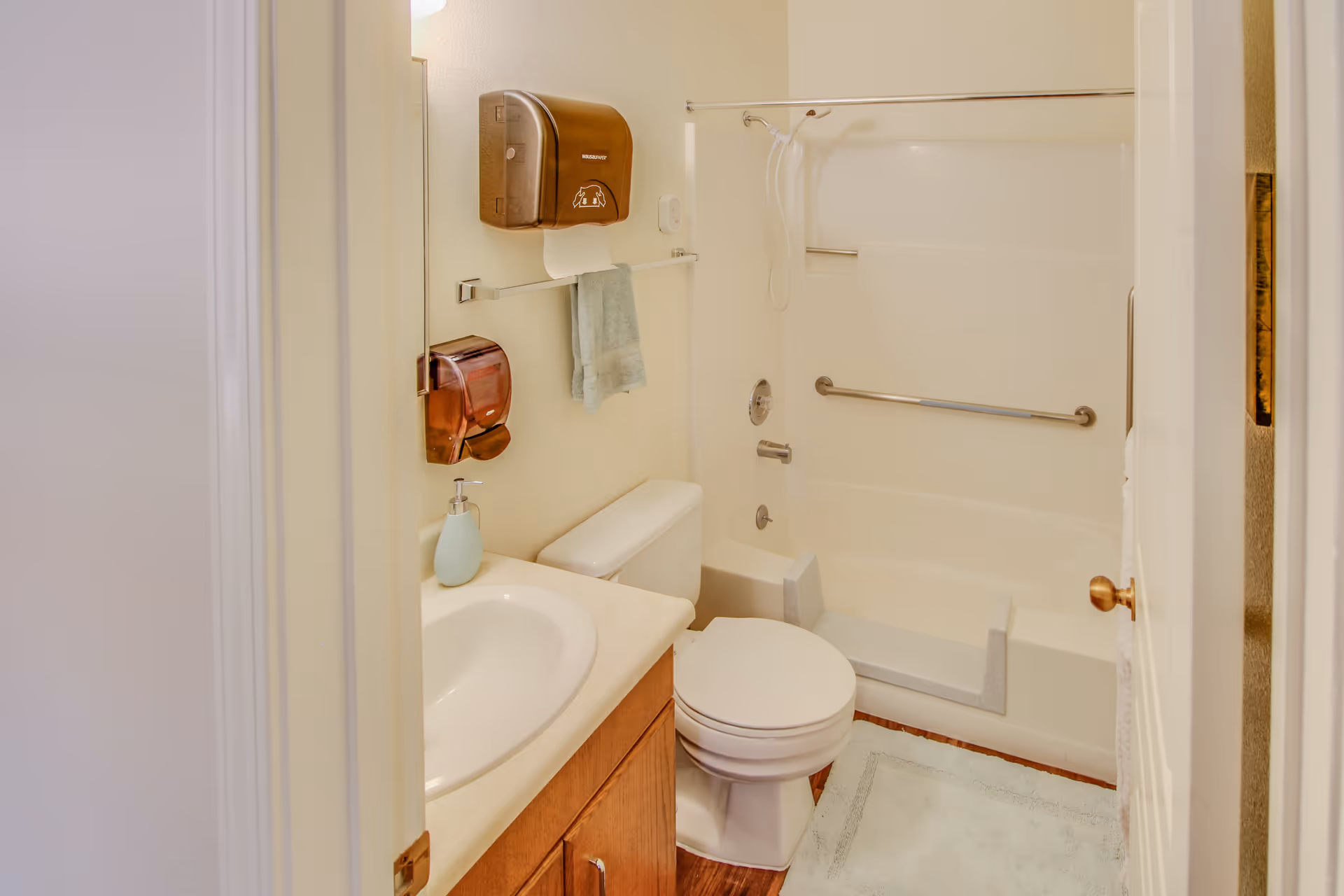 A small bathroom with a white bathtub and shower combination, a toilet, and a sink with a wooden cabinet. There is a towel hanging on a rack above the toilet, a soap dispenser on the sink, and two wall-mounted dispensers above the towel rack. The floor has a light-colored bath mat.