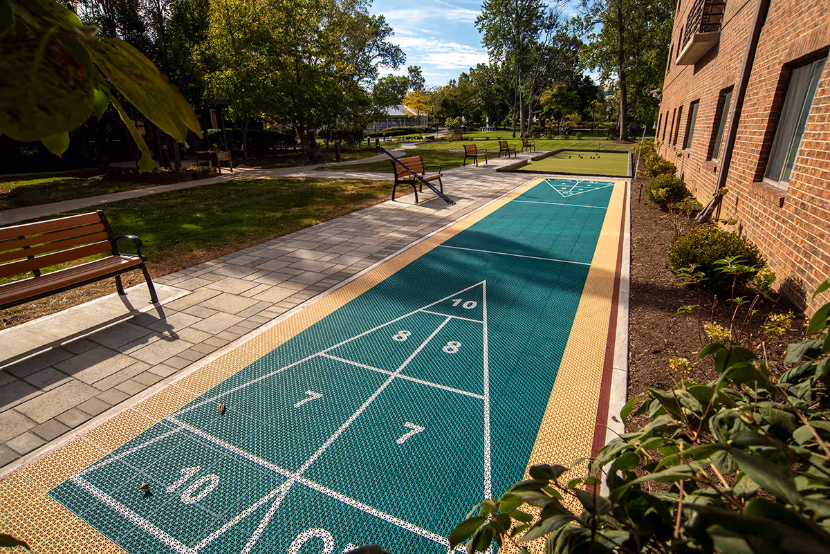 Outdoor shuffleboard court next to a brick building with benches along a paved walkway and green trees in the background under a partly cloudy sky.