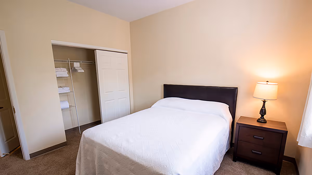 A cozy bedroom featuring a queen-sized bed with a white quilt, a bedside table with a lamp, and a closet with neatly arranged towels.