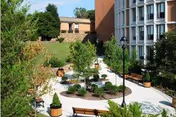 Outdoor courtyard area at Chester County Pocopson Home featuring paved walkways, benches, lamp posts, and landscaped greenery including trees and shrubs, adjacent to a multi-story brick building.