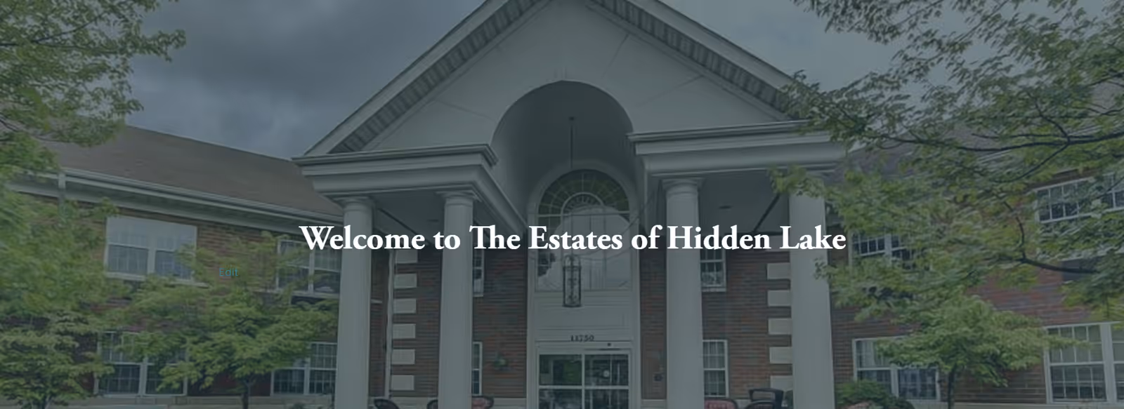 Front exterior view of The Estates of Hidden Lake building featuring a large entrance with white columns, brick facade, and surrounding green trees under a cloudy sky.