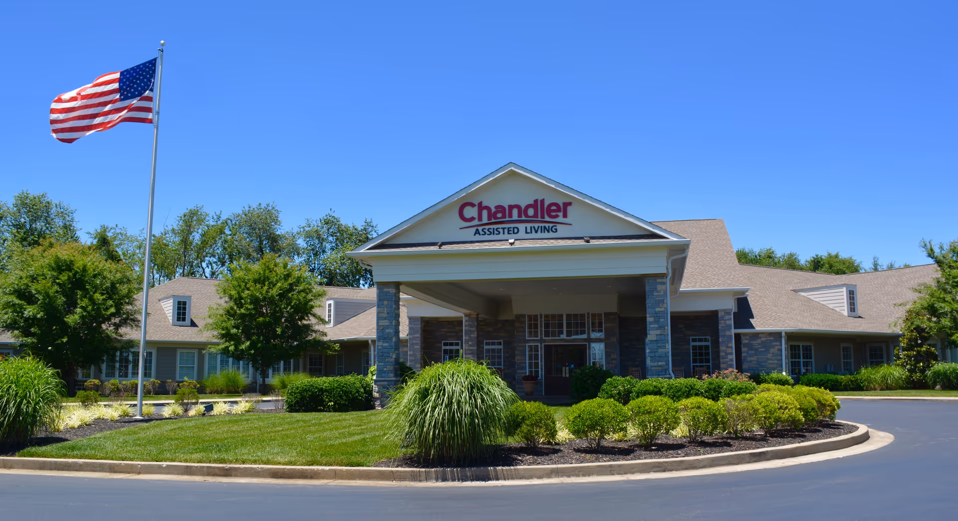 Front entrance of Chandler Assisted Living building with a covered porte-cochère, American flag, and landscaped circular driveway.