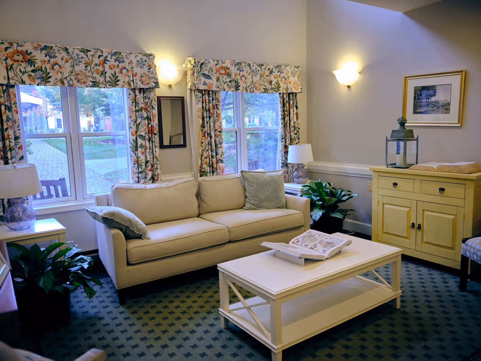 A cozy living room with a beige sofa adorned with two cushions, a white coffee table with an open book on it, floral curtains on two windows, a yellow cabinet with a lantern and a book on top, two table lamps, green plants, and a framed picture on the wall.