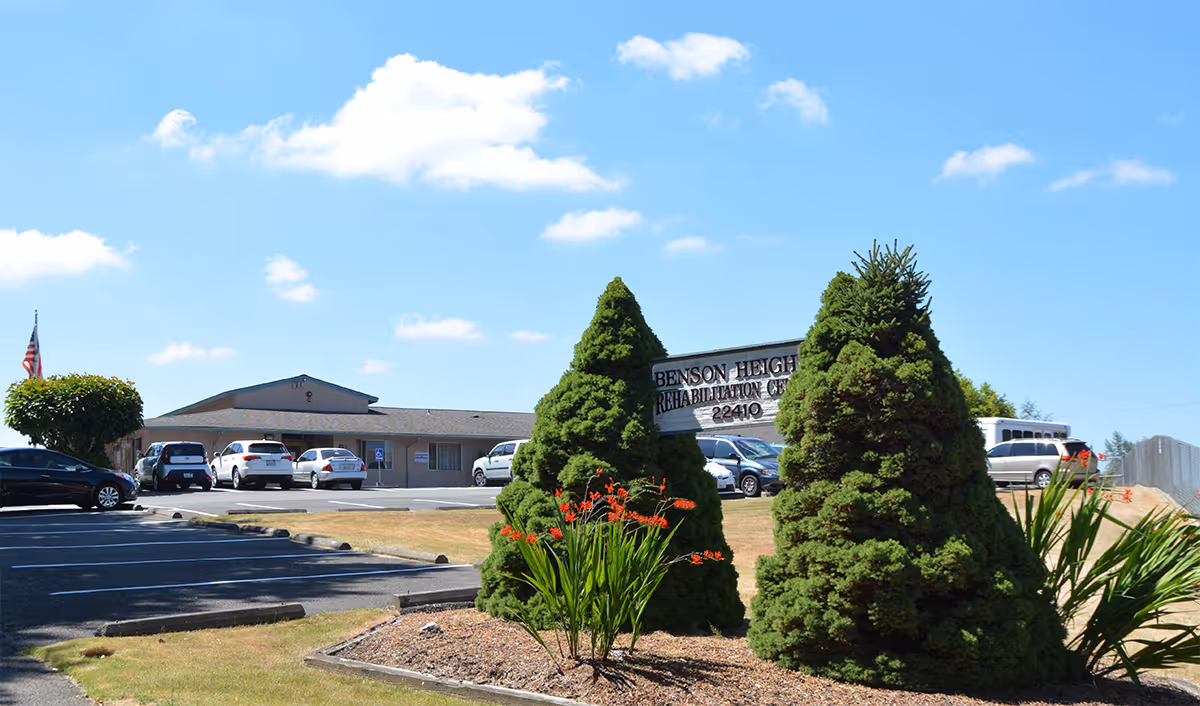 Front exterior of Benson Heights Rehabilitation Center showing the parking lot, landscaped shrubs, and building under a blue sky.