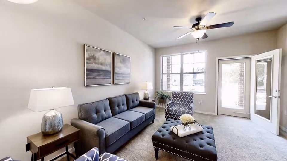 A bright living room with a gray tufted sofa, a matching tufted ottoman with a tray and flowers, a patterned armchair, two table lamps on wooden side tables, two framed landscape paintings on the wall, a ceiling fan with light, and a glass door leading outside.