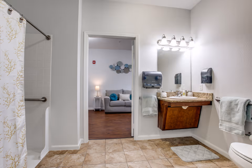 A clean and well-lit bathroom with a tiled floor, a shower with a white and gold patterned curtain, a wall-mounted sink with a granite countertop, a large mirror above the sink, and two towel racks with light blue towels. Through an open door, a living room with a gray sofa, a side table with a lamp, and decorative wall art is visible.