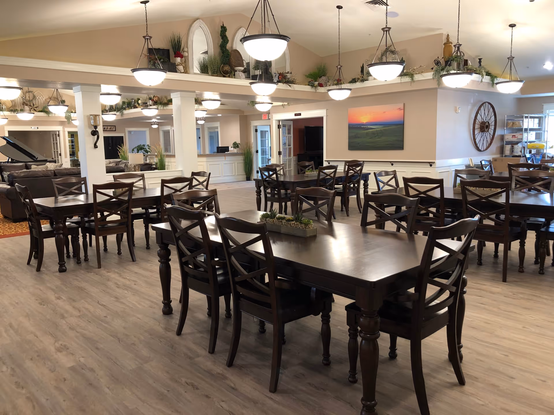 A spacious dining area in an assisted living facility with multiple dark wooden tables and chairs arranged neatly. The room has light-colored walls and wood flooring, with several hanging pendant lights providing illumination. Decorative plants and artwork adorn the walls and shelves, creating a warm and inviting atmosphere.