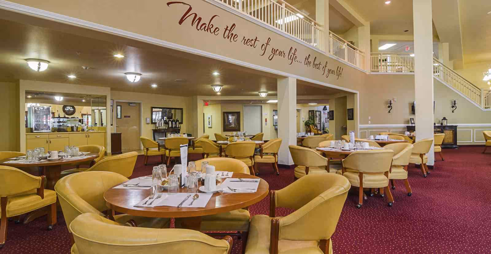 Dining room with round tables set for meals, mustard upholstered chairs, red carpet, and a two-story balcony with wall lettering.