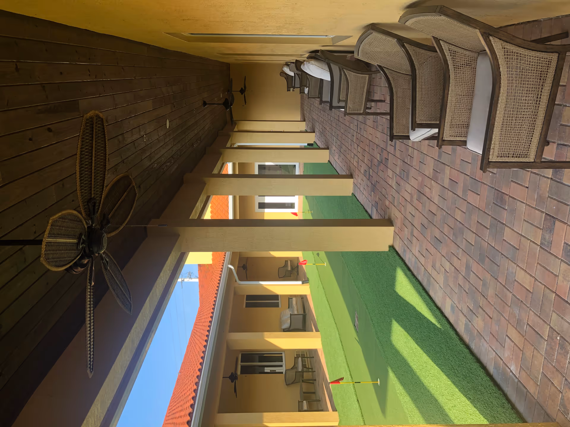 Covered outdoor patio area with cushioned wicker chairs lined up along a brick-paved walkway. Ceiling fans are mounted on the wooden ceiling. Across from the seating area is a small putting green with several red flags, surrounded by yellow walls and a red tile roof under a clear blue sky.