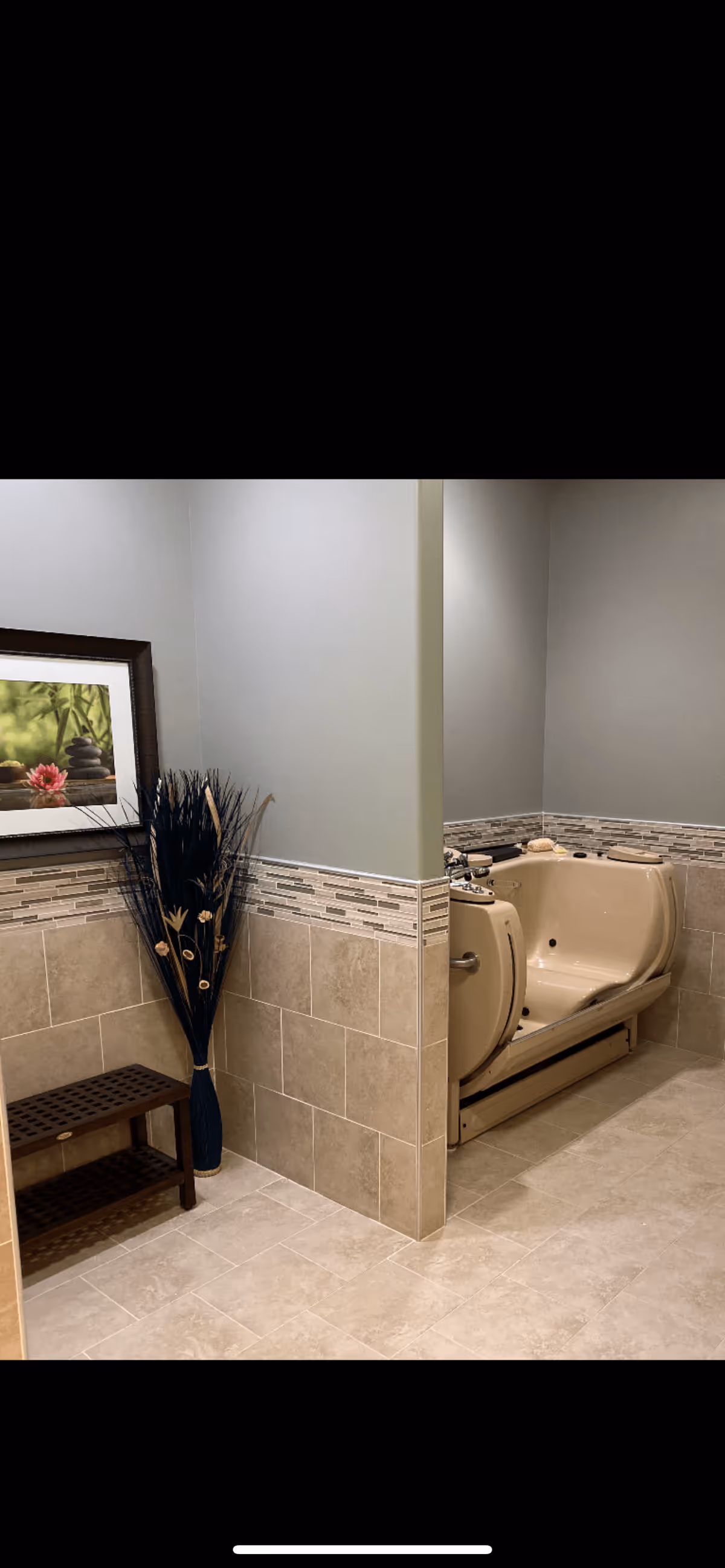 A bathroom area featuring beige tiled floors and walls with a decorative tile border. There is a beige walk-in bathtub with grab bars and a built-in seat. Next to the bathtub is a small wooden bench and a tall vase with decorative dried plants. A framed picture with a nature theme hangs on the wall above the bench.