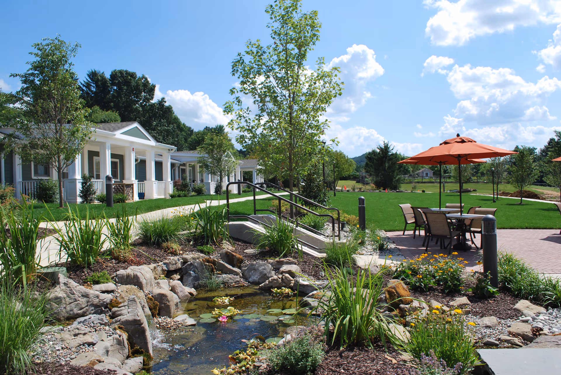 Outdoor area of Heath Village Retirement Community featuring a landscaped garden with a small pond surrounded by rocks and plants. There are paved walkways, green lawns, trees, and patio tables with orange umbrellas. White residential buildings with porches are visible in the background under a blue sky with scattered clouds.