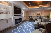 A cozy living room area with a large flat-screen TV mounted above a modern electric fireplace. The room features built-in white cabinetry with shelves displaying decorative items and books. There are several comfortable chairs and a sofa arranged around a blue patterned rug. The ceiling has recessed lighting and a decorative chandelier, and framed artwork is displayed on the walls.