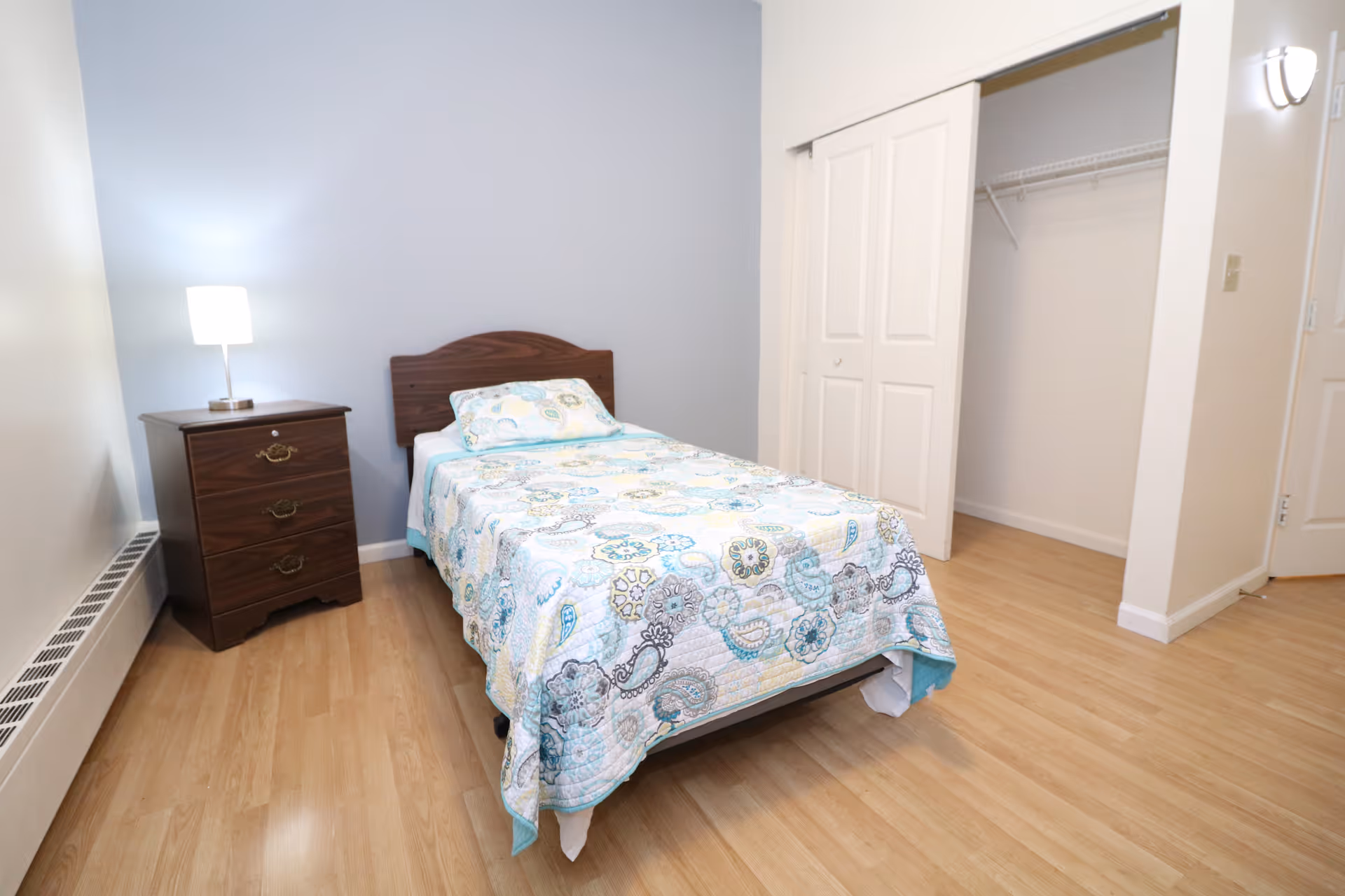 Small furnished bedroom with a single bed, patterned quilt, wooden nightstand with lamp, and an open closet.