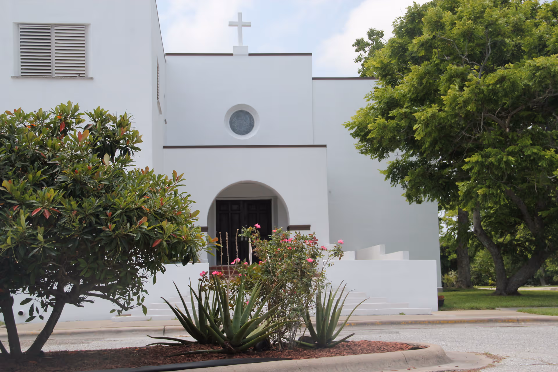 Exterior view of a white building with a cross on top, featuring an arched entrance with dark double doors. In front of the building, there are green shrubs, flowering plants, and trees under a partly cloudy sky.