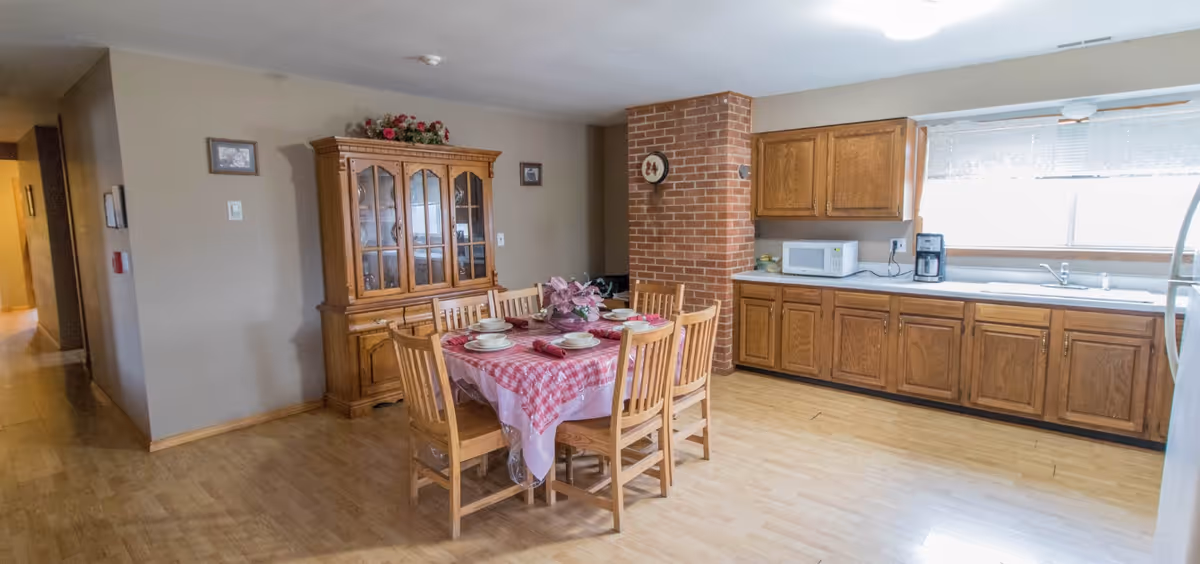 Open dining area with a wooden table set for six, a china cabinet, and an adjacent kitchen with wooden cabinets and a countertop.