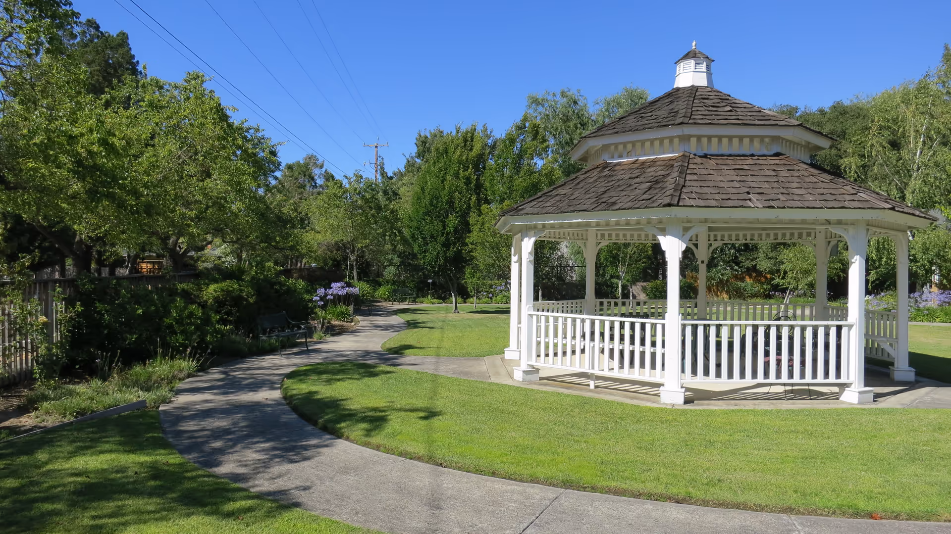 A white wooden gazebo with a shingled roof situated on a well-maintained lawn with a curved concrete pathway leading to it. The area is surrounded by green trees and bushes under a clear blue sky.