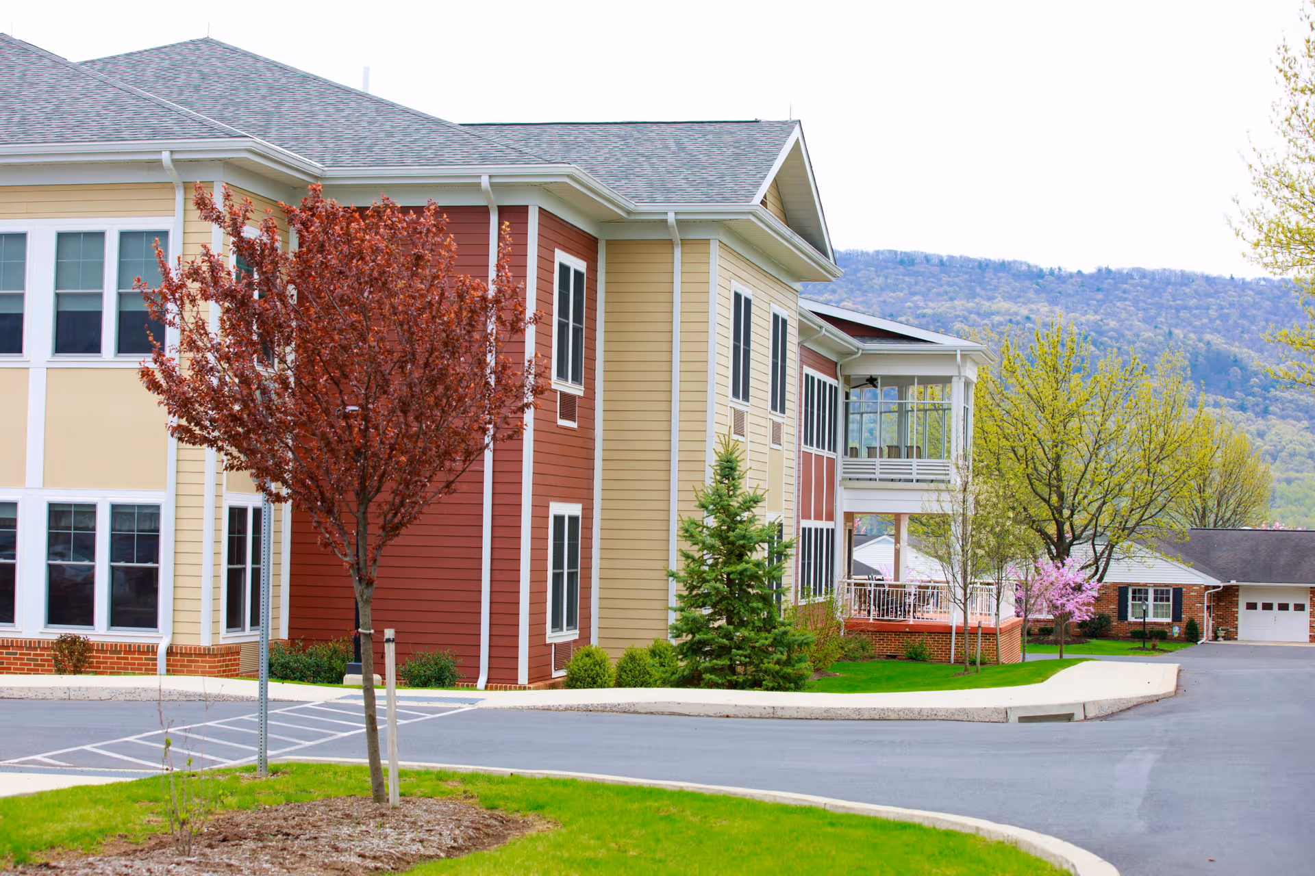 Exterior view of a multi-story building with beige and red siding, surrounded by trees with red and green leaves, a paved driveway, and a mountainous background under a clear sky.