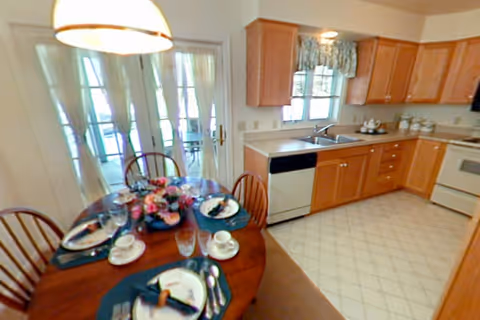 A bright kitchen and dining area with wooden cabinets, a white dishwasher, stove, and sink. A round wooden dining table is set with plates, cups, glasses, and napkins, and a floral centerpiece. French doors with sheer curtains lead to another room.