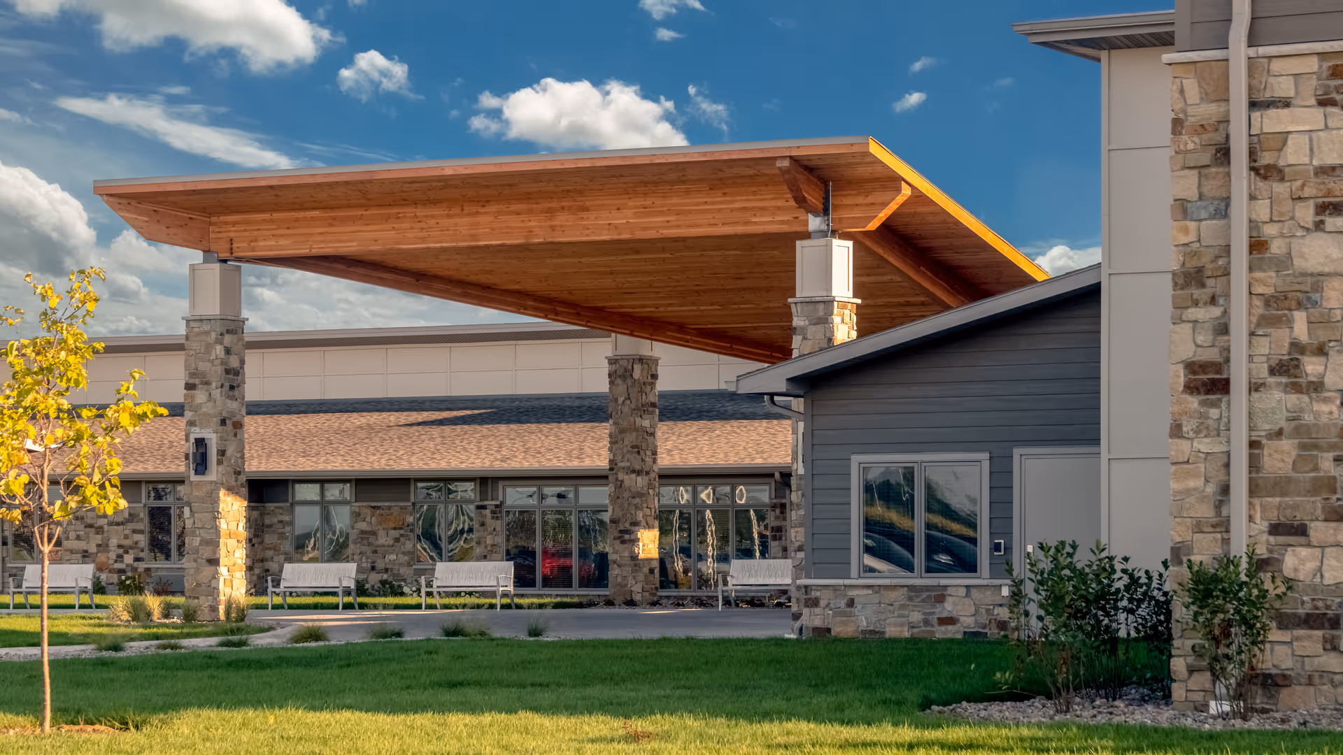 Front entrance of Brookestone Gardens with a large wooden canopy supported by stone pillars, benches, and a landscaped lawn under a blue sky.