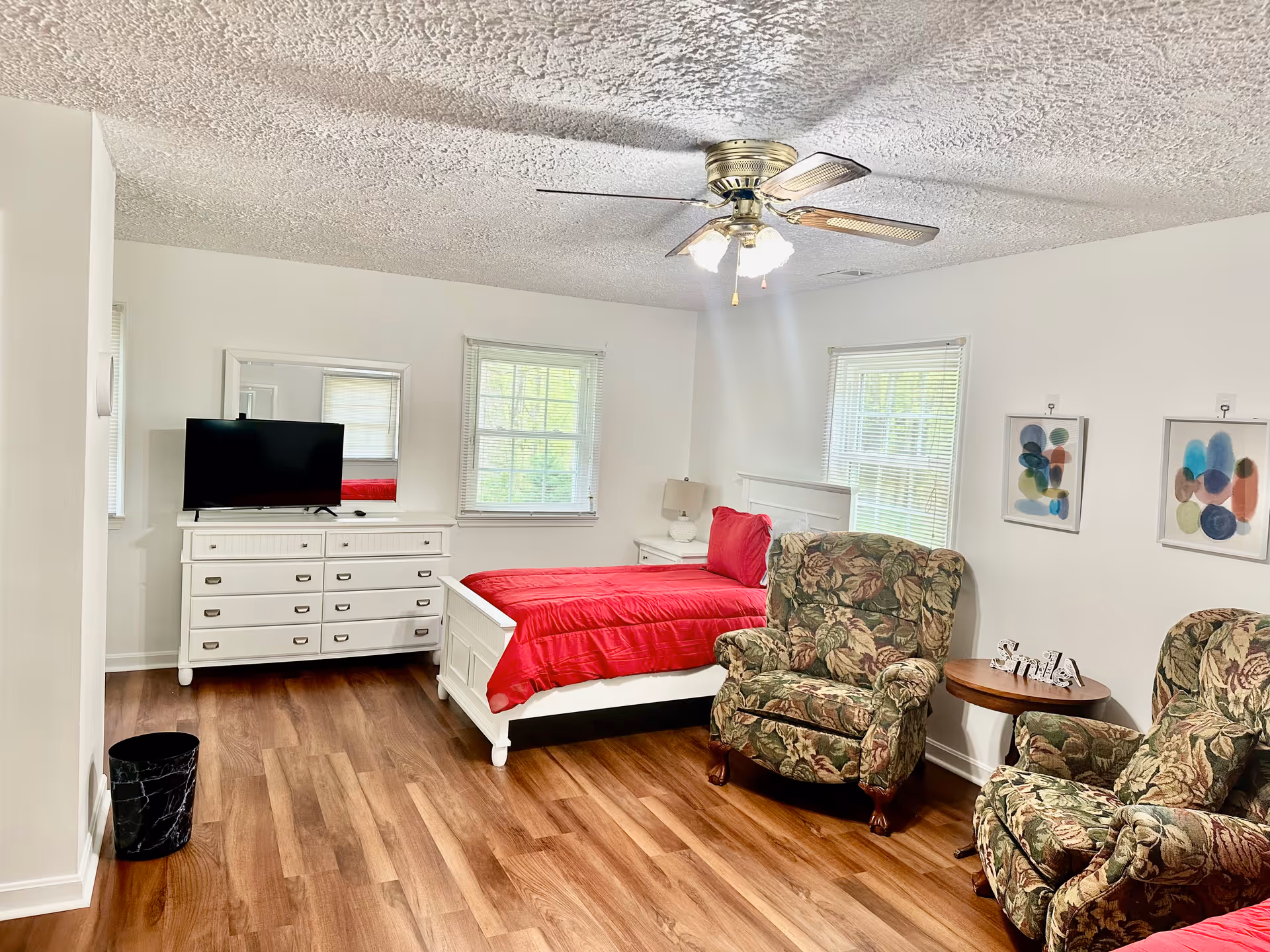 A bright bedroom with white walls and wood flooring featuring a white bed with a red comforter, a white dresser with a TV on top, two floral upholstered armchairs, a small round wooden side table with a decorative 'Smile' sign, two windows with blinds, and two abstract framed artworks on the wall. A ceiling fan with lights is mounted on the textured ceiling.