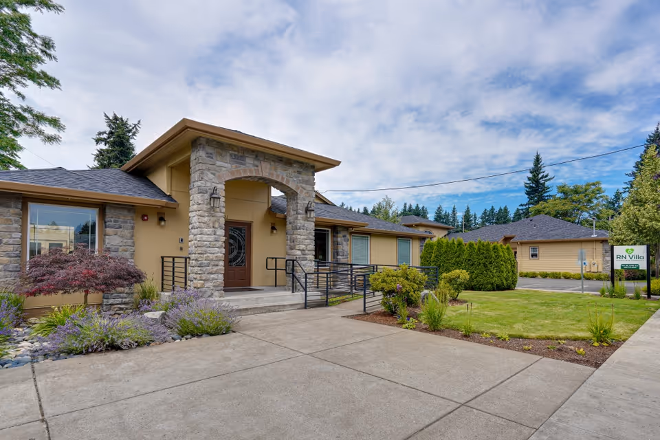 Exterior view of RN Villa Senior Care facility showing a single-story building with stone and beige walls, a covered entrance with stone pillars, a ramp with handrails, landscaped garden with shrubs and flowers, and a sign with the facility name on the right side.