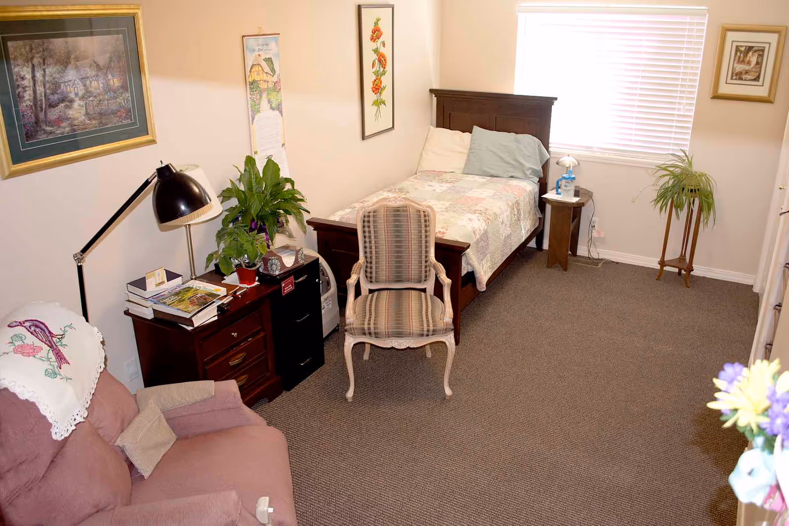 Cozy assisted-living bedroom with a single bed, armchair, side tables, plants and wall artwork.