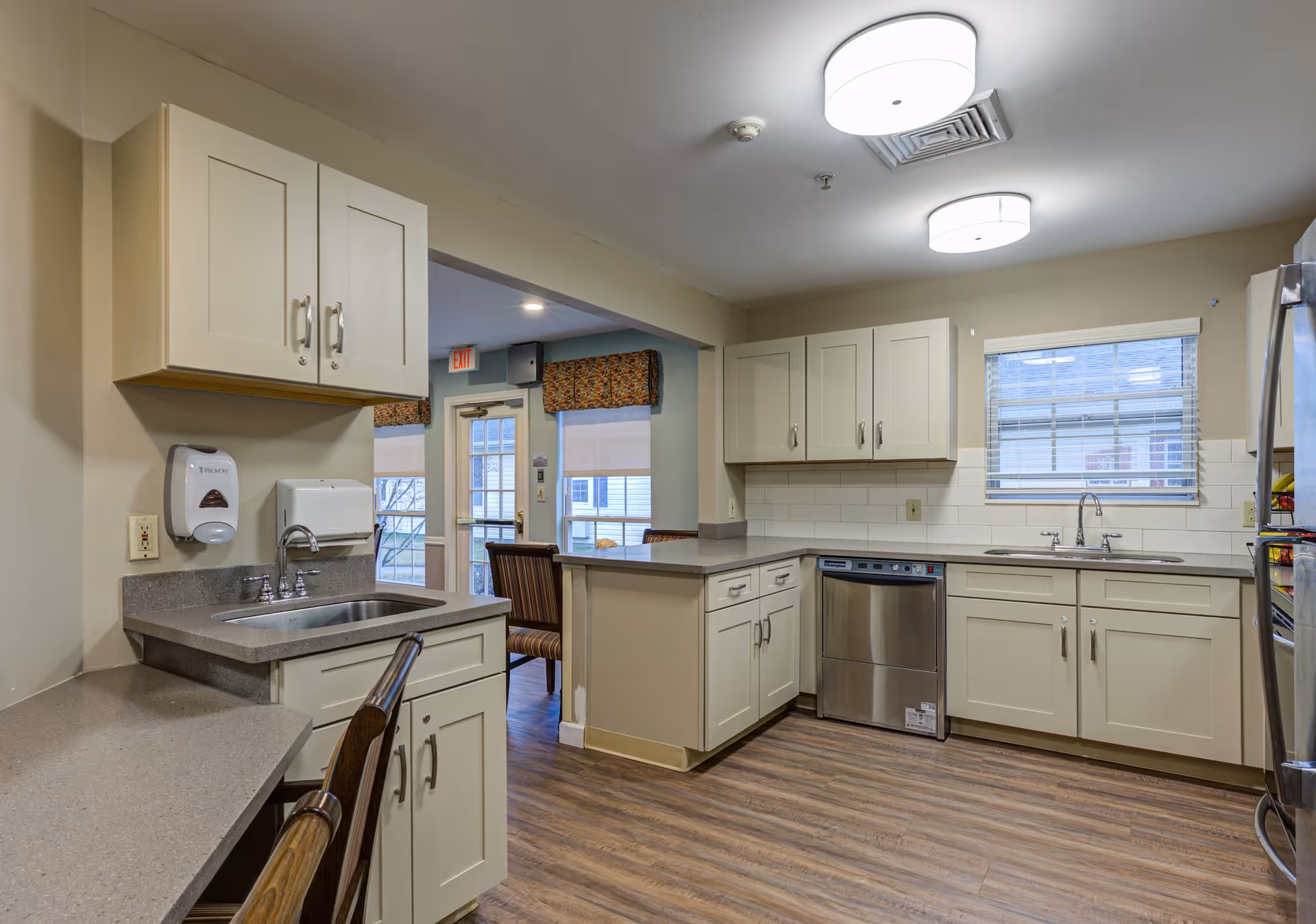 A clean and modern kitchen area with beige cabinets, gray countertops, a stainless steel dishwasher, and a double sink under a window. The floor is wood-patterned, and there are two ceiling lights. In the background, a dining area with chairs and windows is visible.