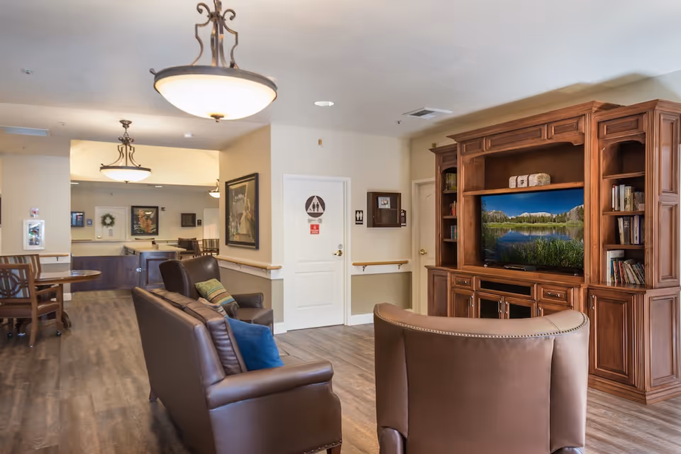 A cozy senior living facility common area with brown leather chairs and sofas arranged around a large wooden entertainment center with a flat-screen TV displaying a nature scene. The room has wood flooring, beige walls, and ceiling lights. In the background, there is a hallway with tables and chairs and a door marked with restroom signs.