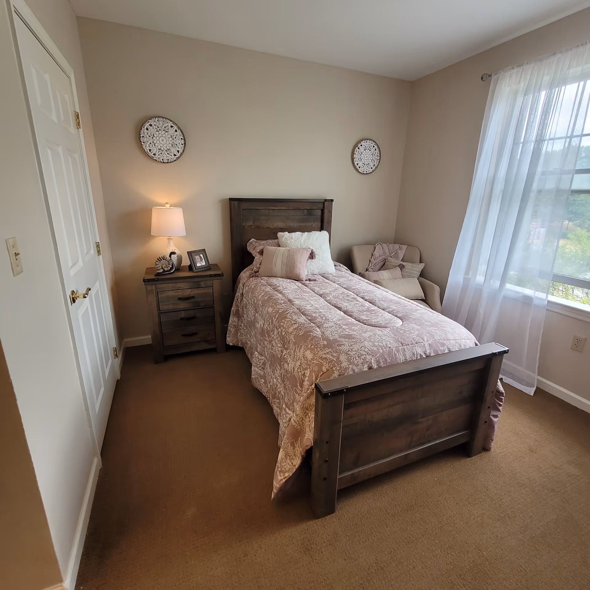 Sunlit bedroom with a wooden bed dressed in a pink patterned comforter, a nightstand with a lamp, and a window with sheer curtains.