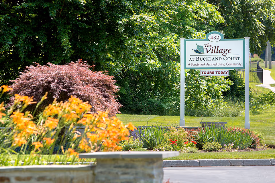 Outdoor view of a landscaped garden area with colorful flowers and shrubs in front of a sign that reads 'The Village at Buckland Court, A Benchmark Assisted Living Community, TOURS TODAY'.